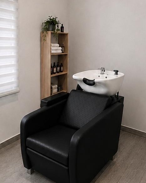 Zona de lavado con sillón negro, lavacabezas y estantería de toallas y productos